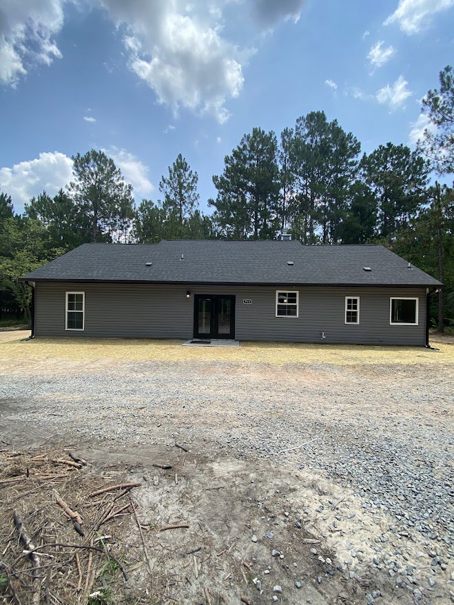 Grey siding house with black front door, large glass doors, gravel and dirt driveway, surrounded by mature trees under cloudy sky