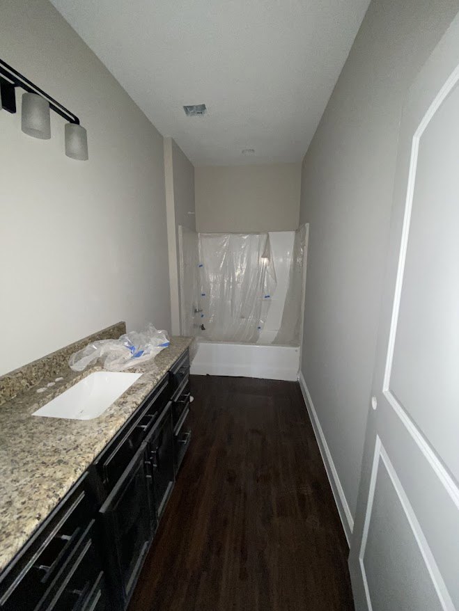 Bathroom with dark wood flooring, freestanding bathtub wrapped in plastic, white countertop sink, light fixture with white shade, neutral tile and plaster finishes