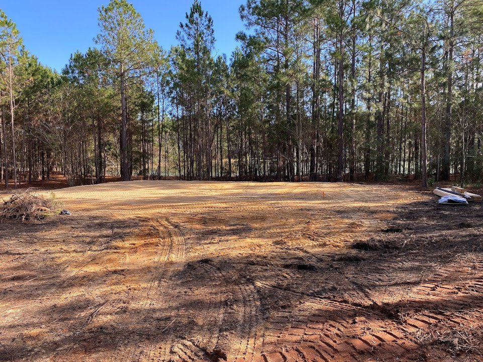 Dirt field with tire tracks bordered by a group of trees under a blue sky, patches of grass scattered across the ground