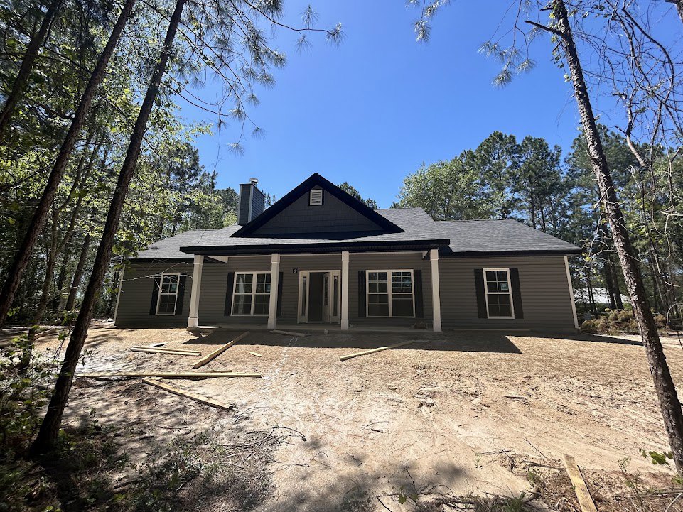 Wood-framed house under construction in a wooded area, featuring a porch, multi-pane windows with white frames, black front door, brick chimney, and dirt yard scattered with