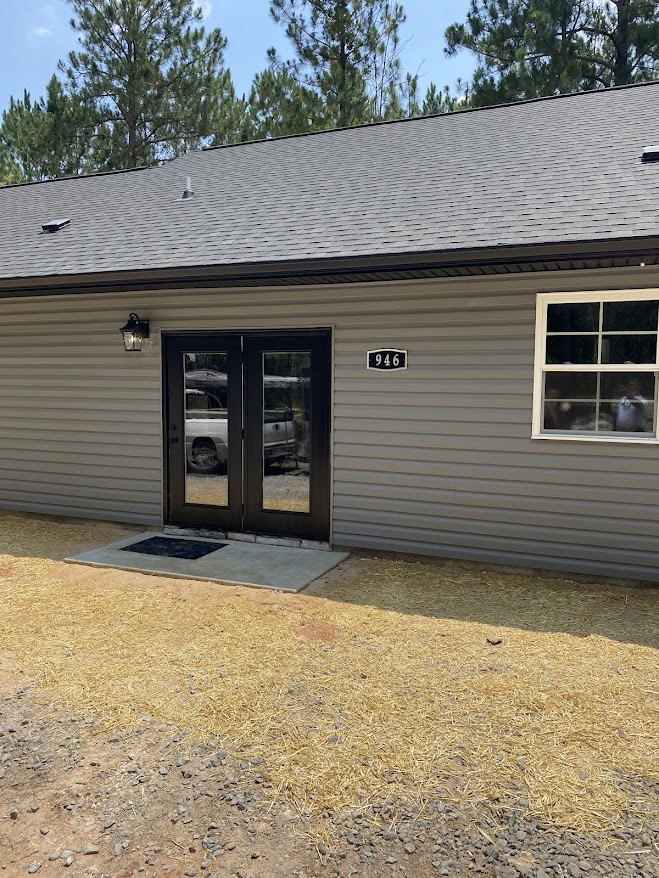 Modern home exterior with double glass doors, large windows, light-colored siding, parked car in driveway, pile of hay on ground, and trees in background under blue sky