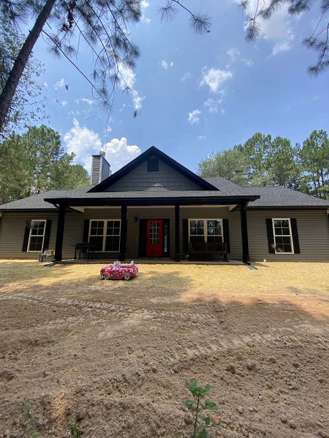 Two-story home with white siding, red front door, multi-pane windows with white frames, tree branches partially visible, toy car on dirt driveway