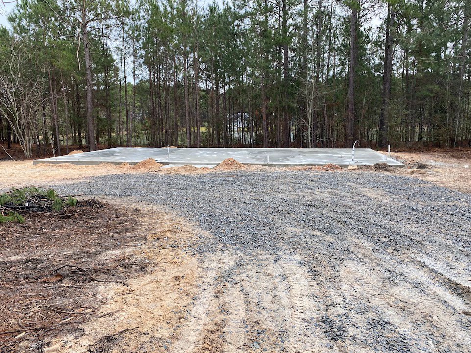 Gravel driveway with visible tire tracks, pile of dirt on concrete, cluster of trees in the background, scattered sticks and soil along the road