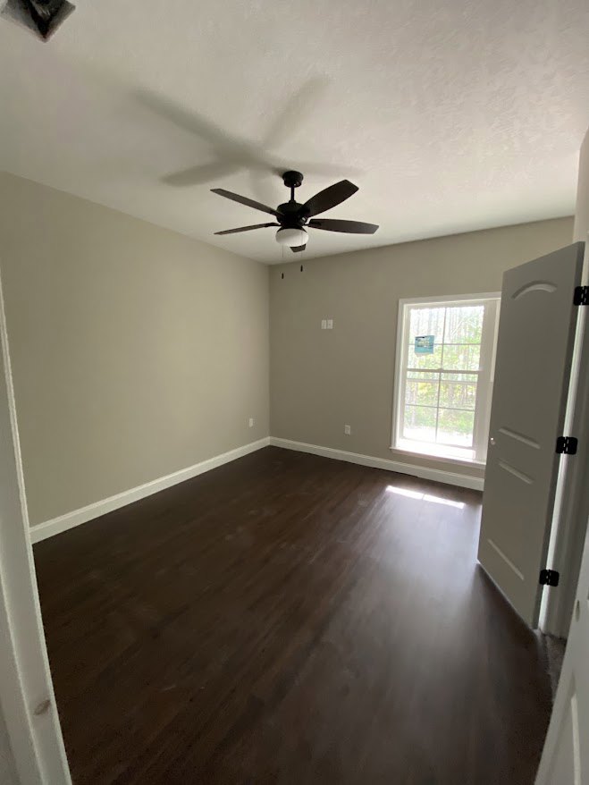 Dark wood flooring, white walls, ceiling fan with light fixture, single window with sign, partial view of door