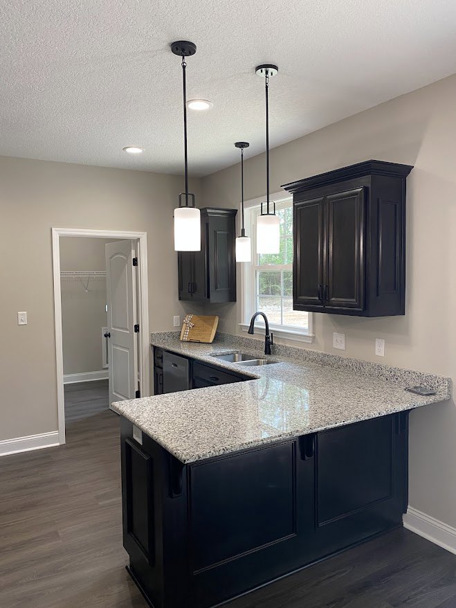 Granite kitchen countertop with undermount sink and chrome faucet, black cabinetry beneath a window, wall-mounted lamp, open door leading to bathroom with tiled floor