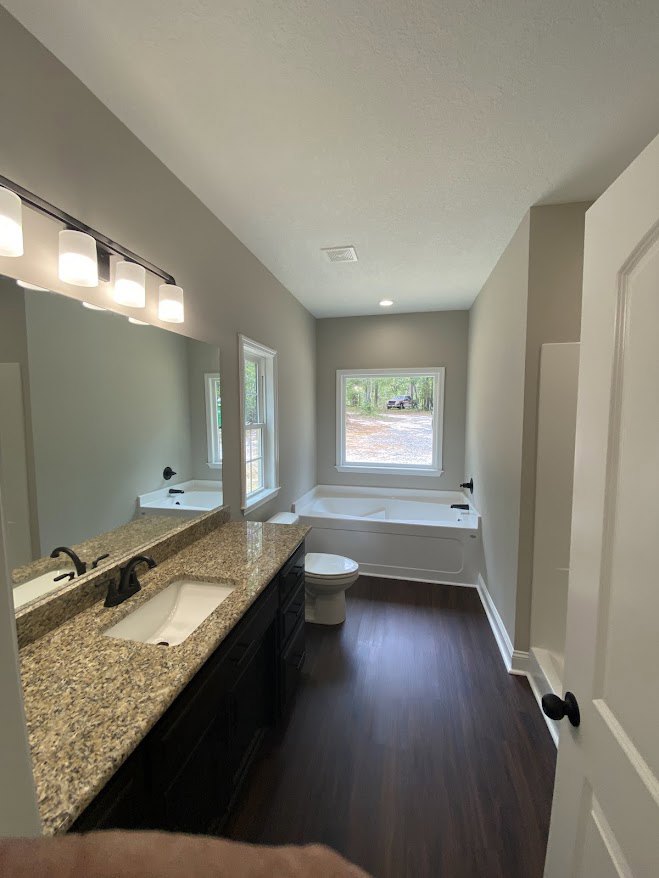Bathroom with marble countertop, white sink and black faucet, freestanding bathtub, white toilet, dark wood floor, white walls, window showing parked car outside