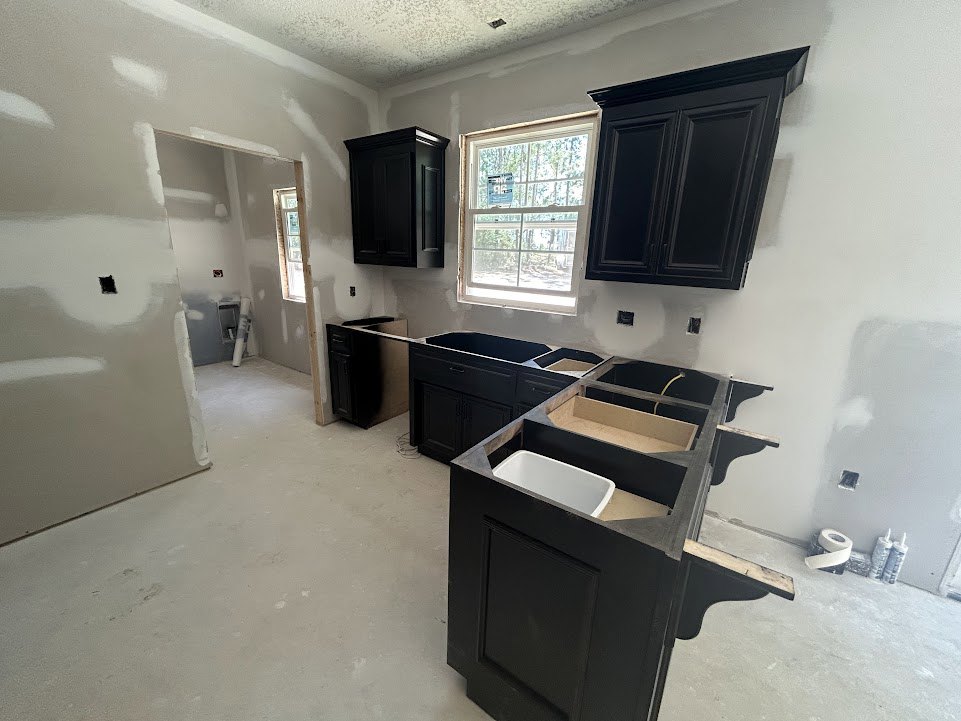 Black kitchen cabinets with matte finish, white tile backsplash, stainless steel sink, light stone countertop, large window with a sign, hardwood flooring, assorted plastic bottles