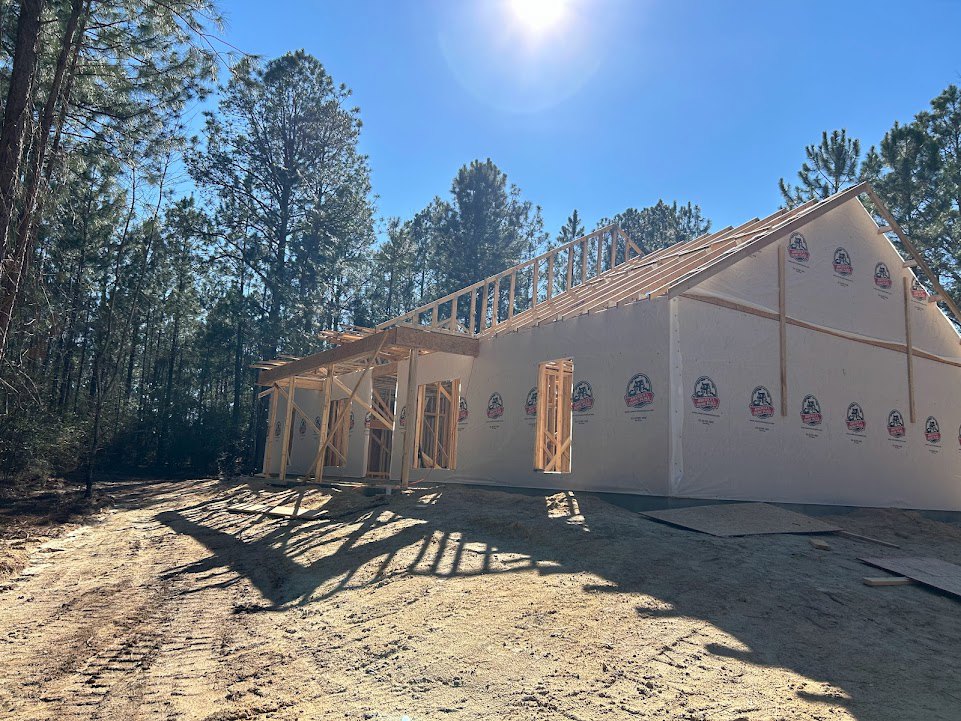 Wood-framed house under construction with exposed beams, sunlit blue sky, and trees in the background