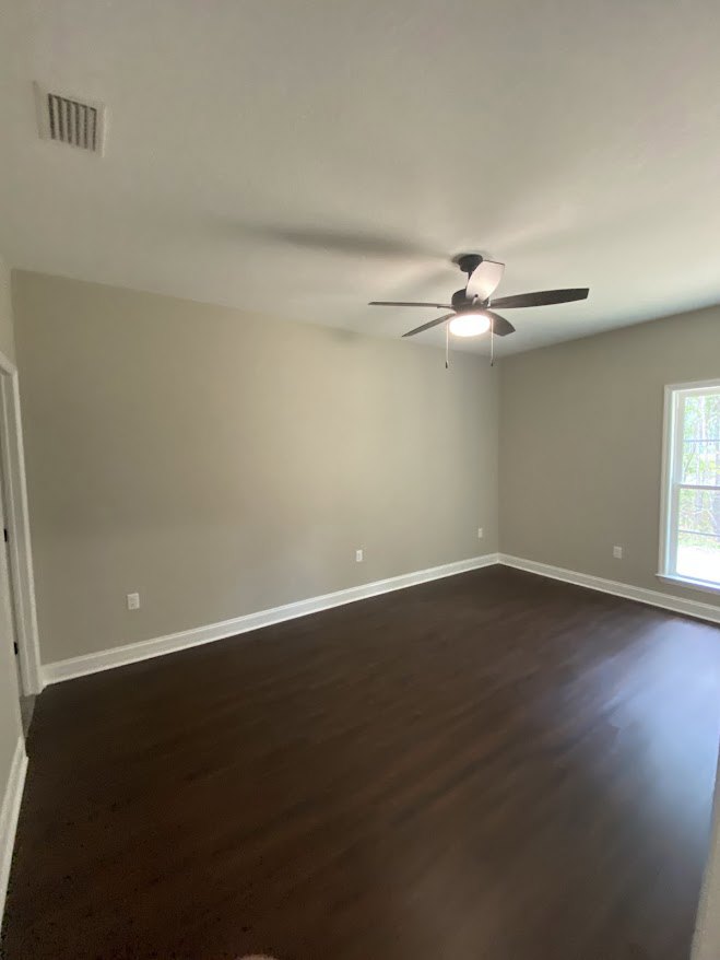 Ceiling fan with illuminated light fixture, dark wood laminate flooring, white framed window, wall vent, smooth plaster walls and ceiling