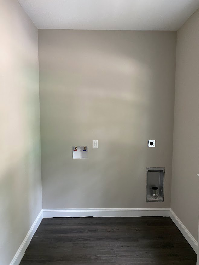 White plaster wall with a recessed metal container, black wood vanity against white background, wood flooring, white electrical outlet, and doorway visible in a modern bathroom.