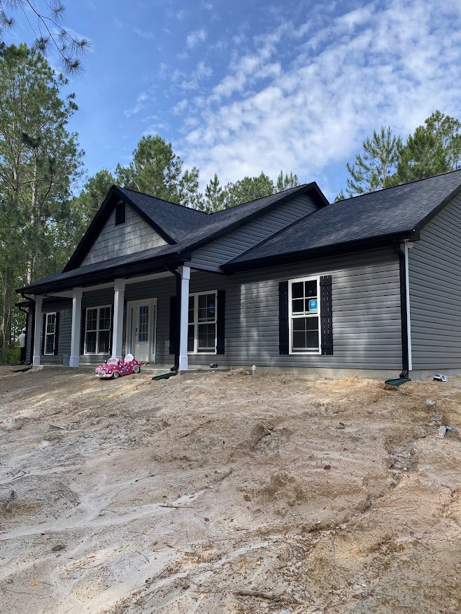Two-story home with white siding, multi-pane windows, and a blue window box; red toy car parked on dirt hill in front; mature trees and cloudy sky in background
