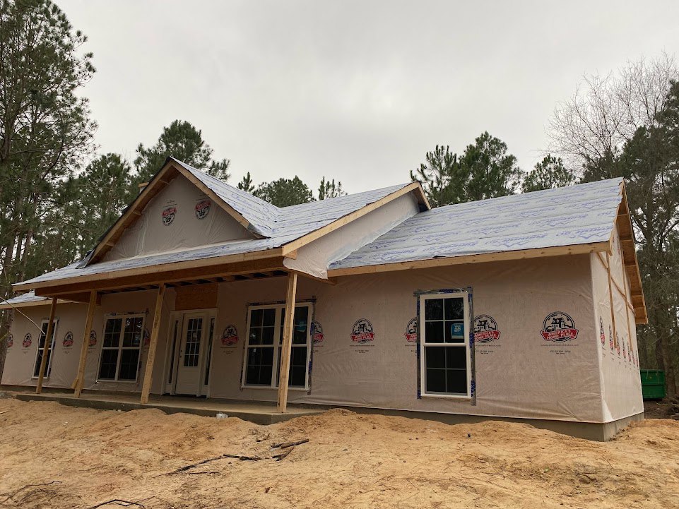 Partially built house with exposed wood framing, white door with glass panes, white-framed window, dirt yard scattered with sticks, trees in background
