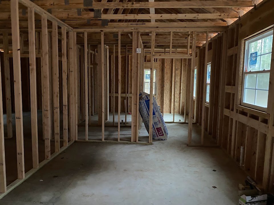 Unfinished interior room with exposed wooden beams, concrete floor, large roll of white paper on a wooden frame, blue sign taped to window, and white and blue bag of material.