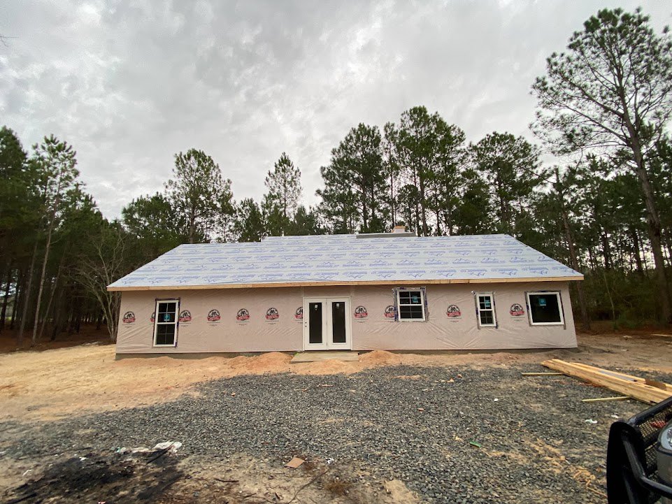 Partially built house with exposed framing, white double door featuring black glass panels, gravel-covered ground, window displaying a blue sign, surrounding trees in the
