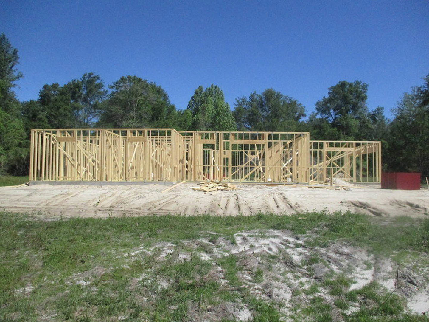 Wood-framed house under construction with exposed beams, grassy foreground, pile of lumber, trees and blue sky in background