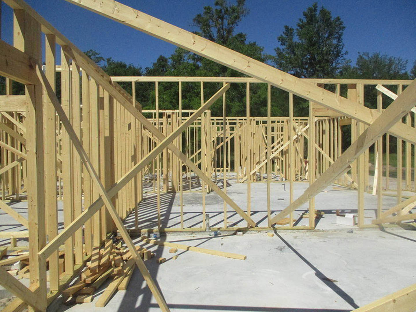 Framed house under construction with exposed wooden beams, concrete foundation, scattered lumber, and leafy tree in background