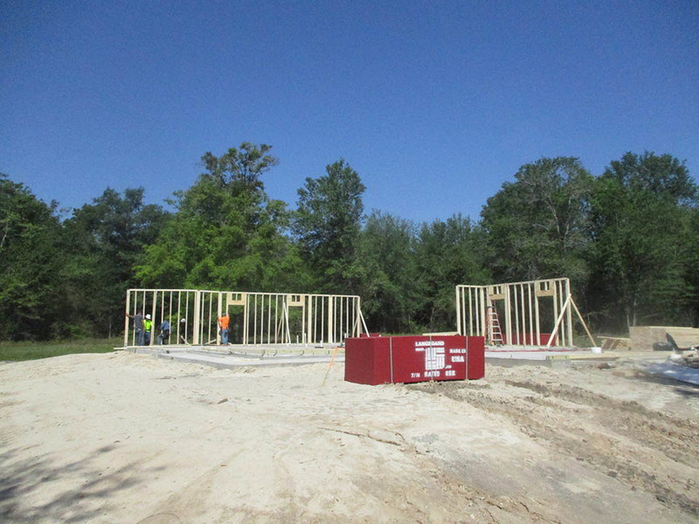 Construction site with sandy ground, a ladder leaning against a wall, a man standing near a metal gate, red box with white text placed in the sand, fenced perimeter, and trees in