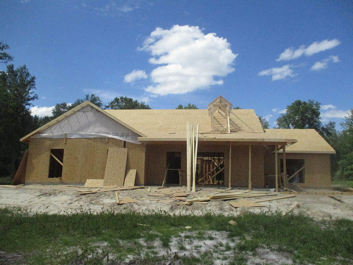 Partially built wooden house with exposed framing, scattered wood planks, unfinished roof, and cloudy sky overhead