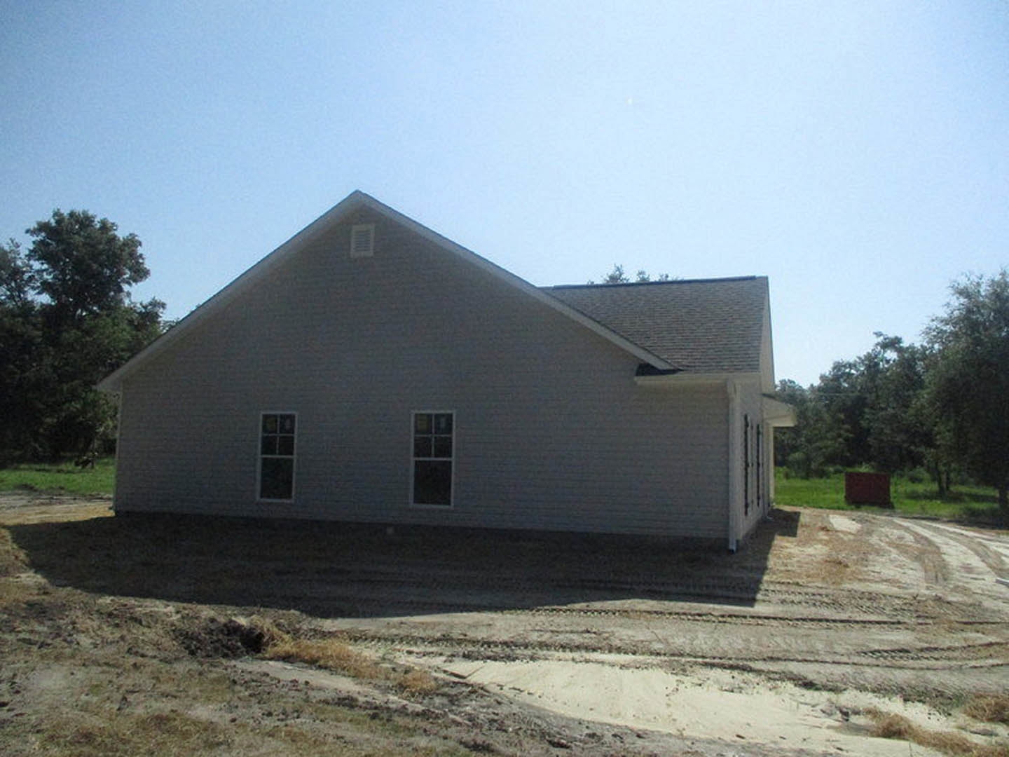 Brown-sided house with white-trimmed windows, surrounded by dirt lot and mature trees in the background