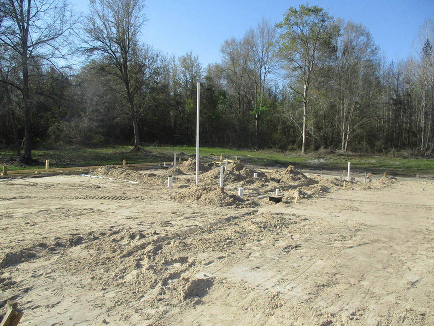 Dirt field with scattered piles of soil, white poles, leafless tree, and group of trees in the background under a clear sky