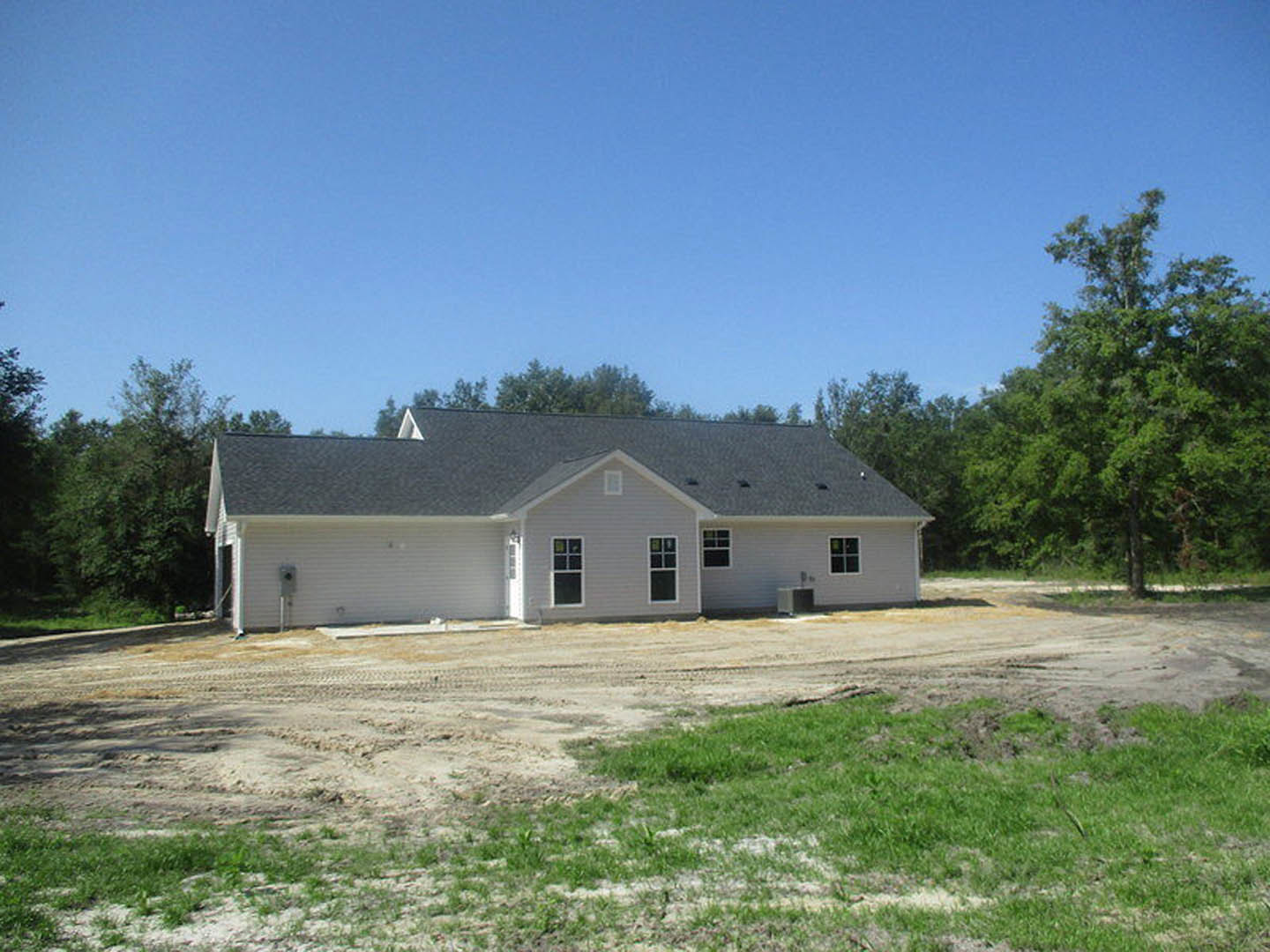 Partially built house with grey roof and visible windows, surrounded by grassy field and leafy tree under blue sky with scattered clouds, dirt patch in front of structure.