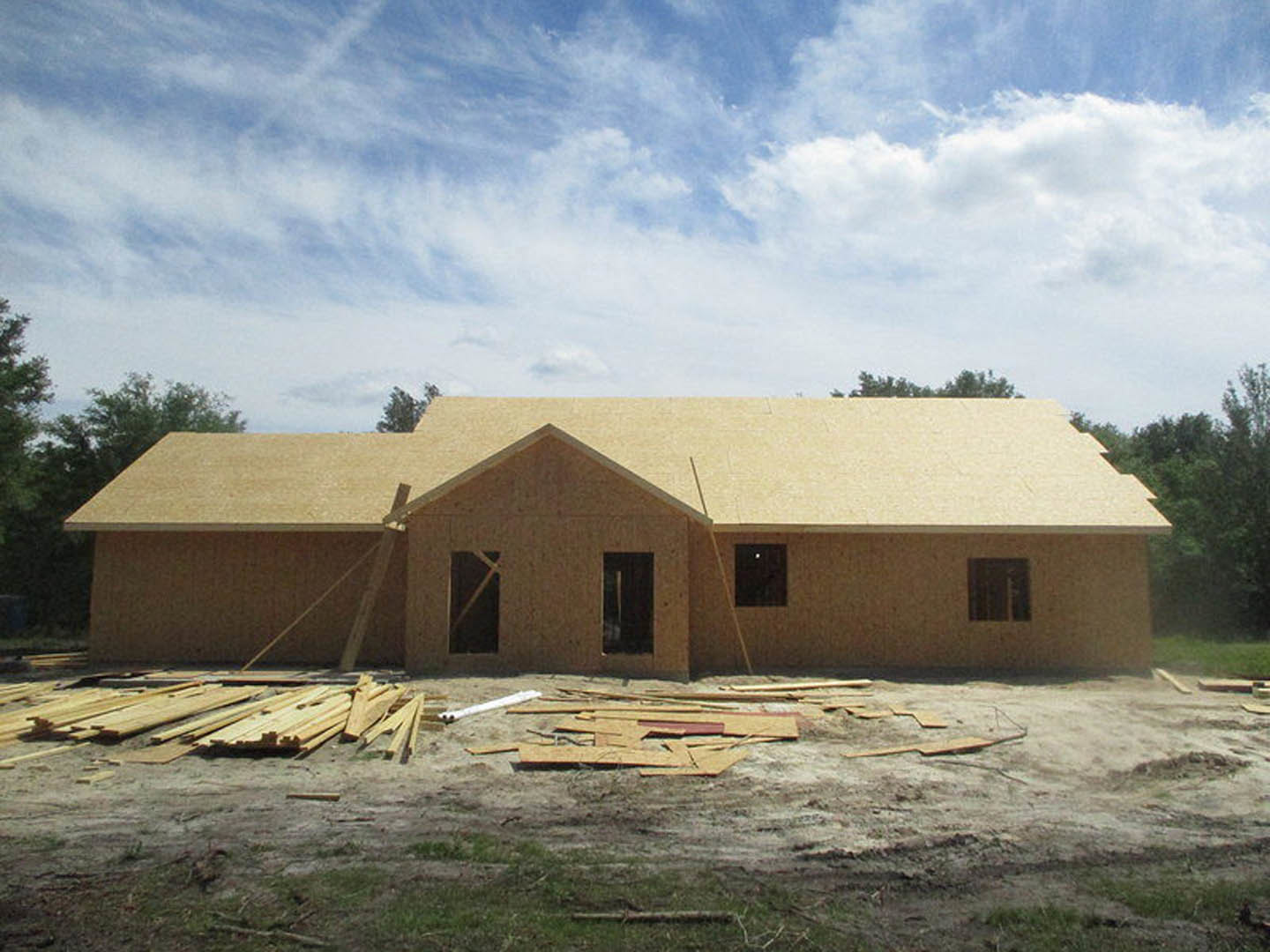 Framed house under construction with exposed wood planks, partially completed roof, pile of lumber on dirt lot, blue sky and scattered clouds in background