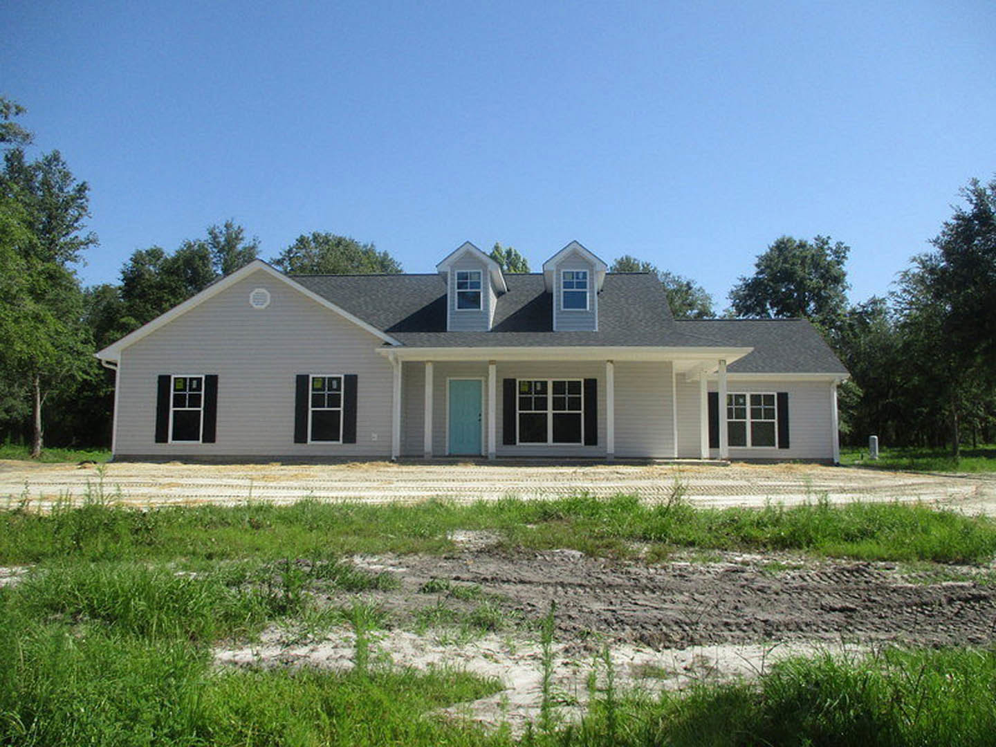 Partially constructed house with blue front door, white-framed window, unfinished porch, dirt patch in grassy yard, and exposed building materials