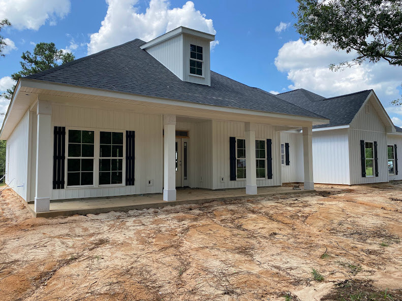 White house with black shutters and multi-pane windows, white columns supporting a porch, dirt ground in front, cloudy sky overhead.