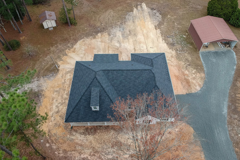 Modern home exterior with light-colored siding, large windows, and a sloped roof, surrounded by bare dirt and mature trees