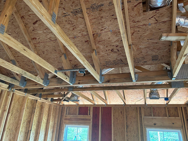 Exposed wooden framing and beams in a partially constructed house, with a window opening and ladder outside, unfinished walls and ceiling visible