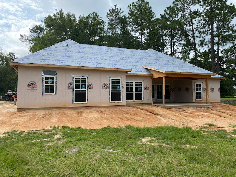 Partially built house with exposed framing, blue roof panels, white door with glass panes, surrounded by brown dirt and patches of grass, mature trees in the background