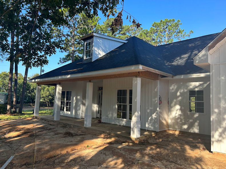 White siding house with black shingle roof, rectangular white-framed windows, covered porch, dirt yard, and mature trees in the background