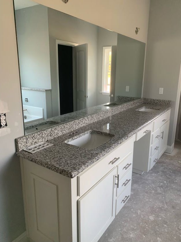 Bathroom with expansive mirror above white sink and gray granite countertop, white cabinetry with silver handles, bathtub with metal shower rod, tiled walls and floor.