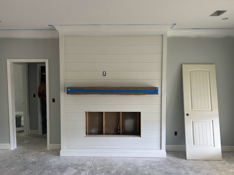 Living room with unfinished fireplace, white plaster walls, black pipe and yellow wire exposed, white door with central hole, floating shelf with blue painter’s tape, man standing