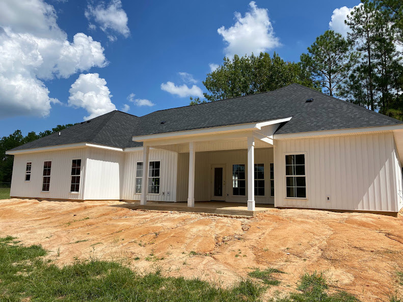 White siding house with black shingle roof, white-framed windows, dirt patch in front yard, green leafy trees and blue sky in background, sunlight casting shadow on exterior wall.
