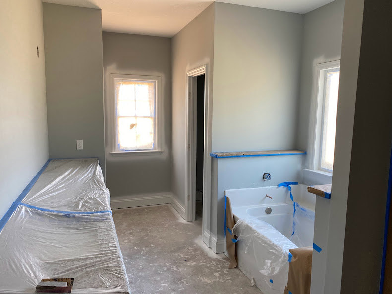 Bedroom with a plastic-wrapped mattress and bathtub, sunlight streaming through window, tiled floor, plaster walls, plumbing fixtures visible