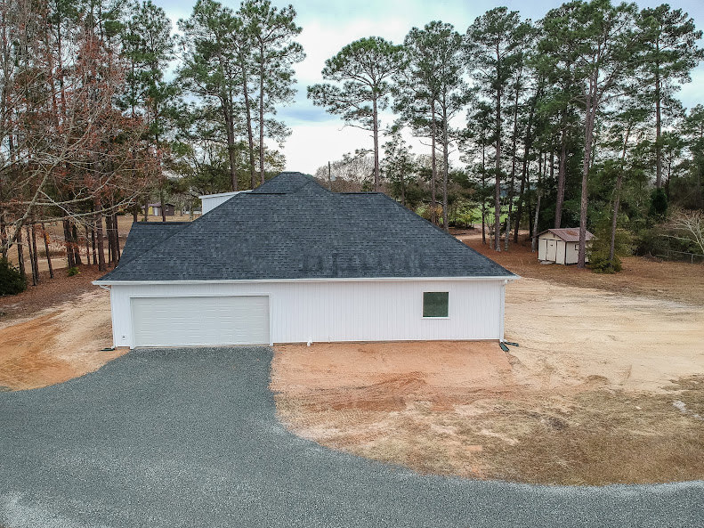 Modern two-story home with gray shingle roof, wide concrete driveway, and mature trees surrounding landscaped front yard
