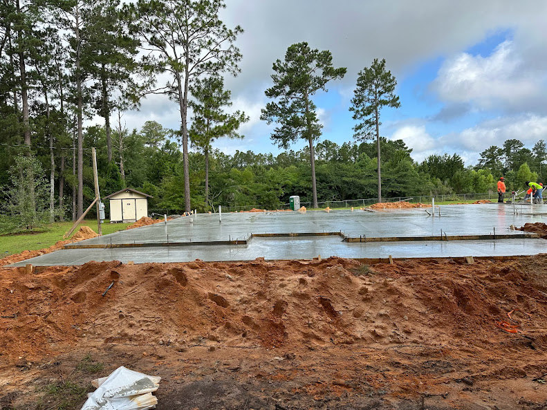 Concrete foundation slab surrounded by tall green trees, blue sky overhead, white shed with brown roof in background