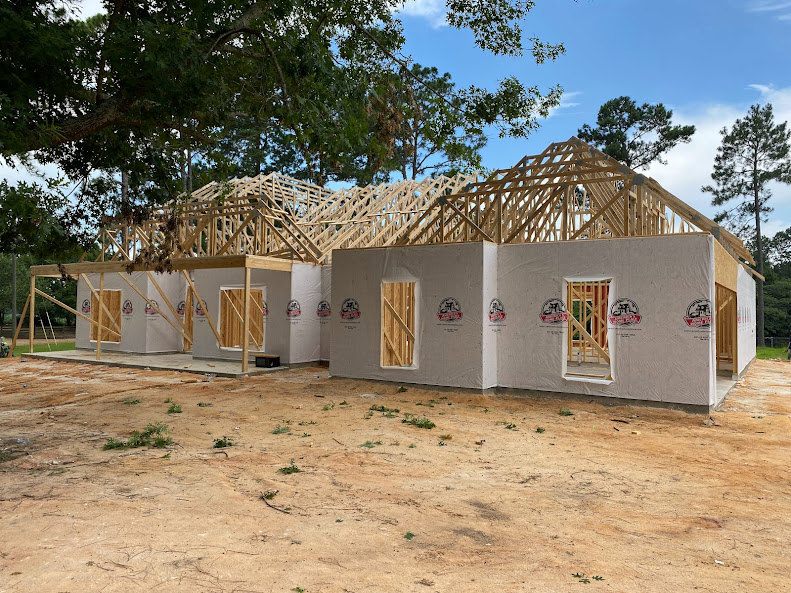 Framed wood structure of a house under construction, exposed beams and unfinished walls, tree nearby, dirt ground with scattered plants, open window cutout, light-colored framing