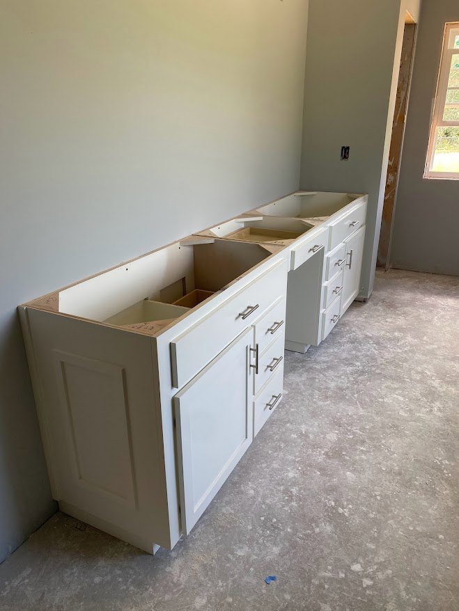 White kitchen cabinets with multiple drawers, white countertop, window with white frame, brown door frame with white trim, light-colored walls and flooring