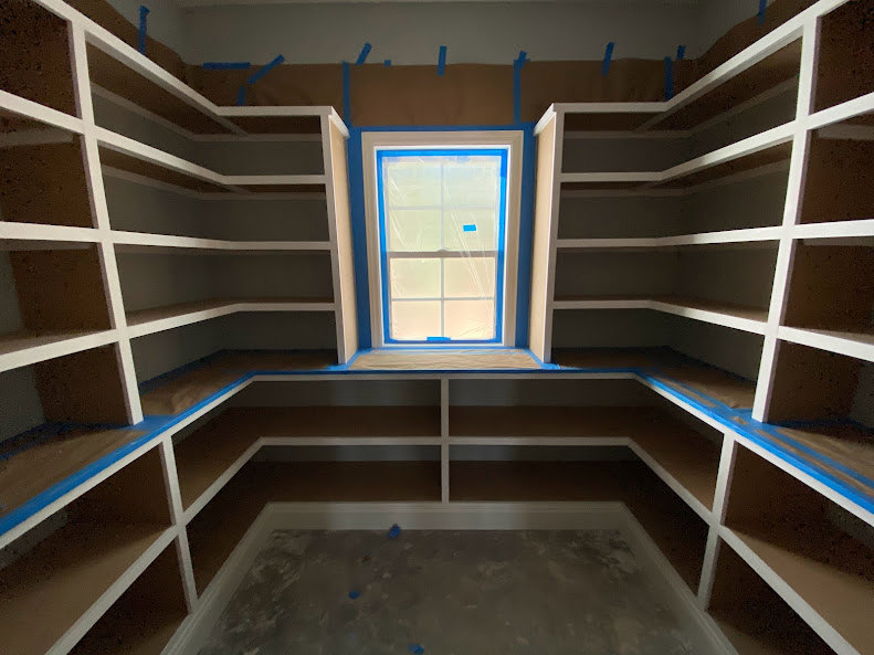 Built-in shelves along a white wall, grey flooring with scattered blue objects, window partially covered with plastic tape, staircase visible in background.