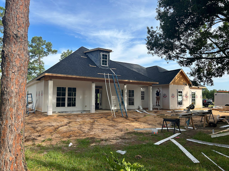 Two-story house under construction with exposed wooden framing, multiple ladders leaning against the structure, unfinished windows, and a tree trunk in the foreground.