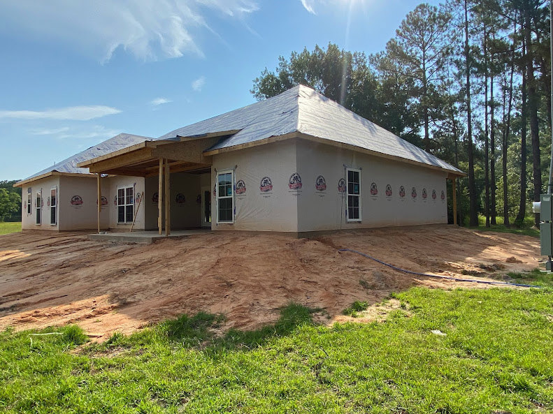 Wood-framed house under construction on grassy lot, surrounded by mature trees, blue sky with scattered white clouds overhead