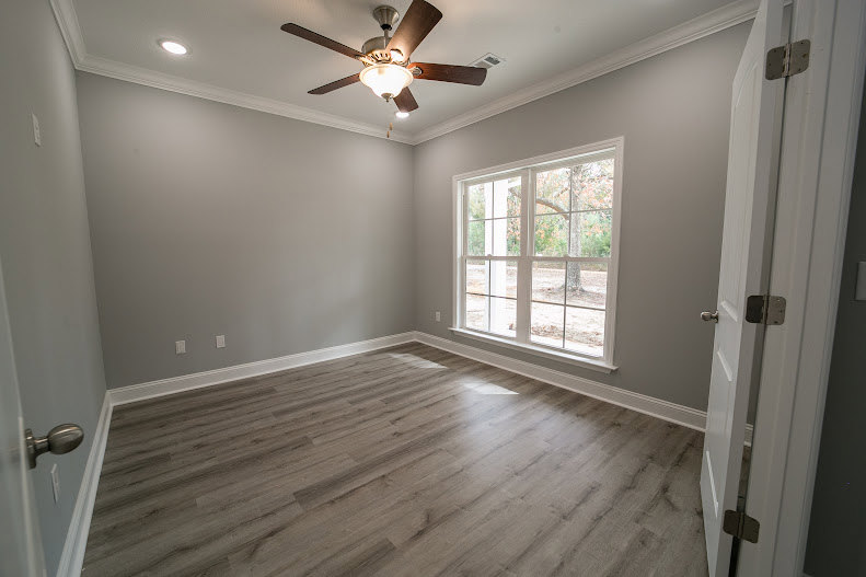 Wood flooring, white plaster walls, ceiling fan with light fixture, large window with trim molding, natural light illuminating interior space