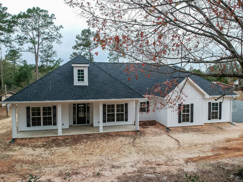 White siding house with black shingle roof, multiple windows, surrounded by trees and lawn under blue sky