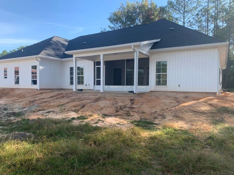 White siding house with black roof, front porch, white door, rectangular windows, grassy yard with scattered rocks, trees in background under blue sky