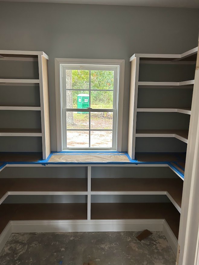 Built-in white shelving along a grey wall, large window with daylight streaming in, white ladder leaning nearby, green container visible outside.