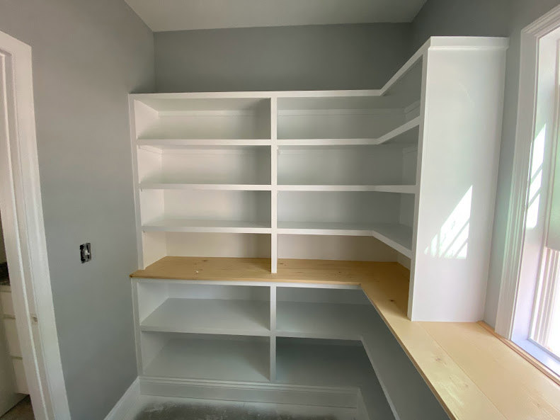 White shelving unit with wooden top against plaster wall, white curtain with black stripe beside window, sunlight streaming into room, hardwood flooring visible.