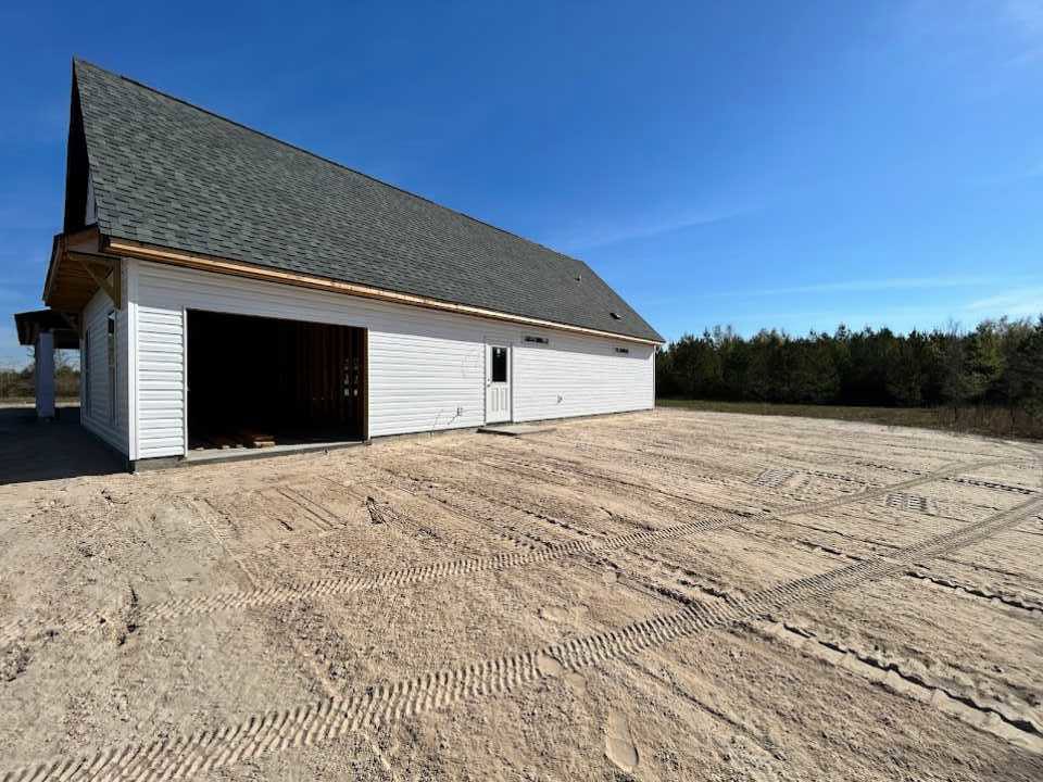 Modern house with attached garage, unfinished dirt yard with tire tracks, man standing inside garage, light-colored roof, distant trees under partly cloudy sky