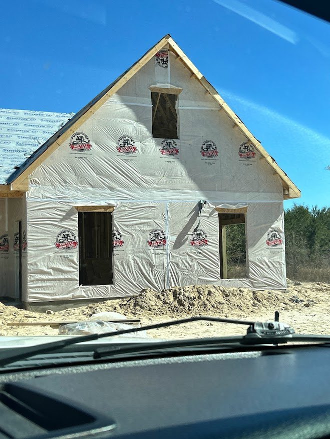 Partially built house with exposed plywood walls, finished shingle roof, plastic sheeting covering windows, parked car in driveway, leafy tree and blue sky in background